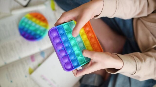 Teenage girl playing with rainbow pop-it fidget toy while studying at home. Teen kid with trendy stress and anxiety relief fidgeting game. Popping the dimples of sensory silicone toy.