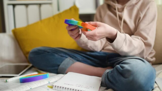 Teenage girl playing with rainbow pop-it fidget toy while studying at home. Teen kid with trendy stress and anxiety relief fidgeting game. Popping the dimples of sensory silicone toy.