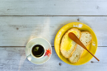Ghee butter on sliced bread, with wooden spatula, in a yellow plate next to a cup of coffee, on a white wooden table.