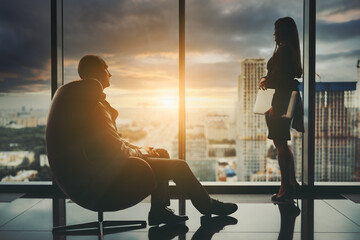 Silhouettes of two business persons in front of a window of a skyscraper observing a stunning sunset an urban landscape after a hard workday, with selective focus on the businessman in an armchair