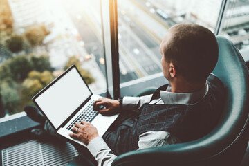 View of a businessman with a laptop on his knees with a white screen mock-up, sitting on an armchair and looking down on the highroad from the window of a luxury business skyscraper, selective focus