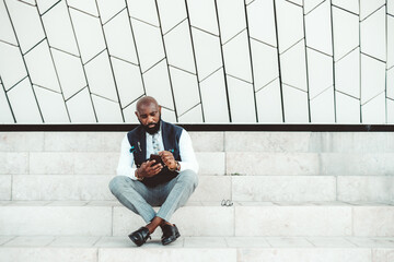 A fashionable bald black senior entrepreneur with a beard and in an elegant suit is sitting on a marble step on the street, in front of a patterned wall and texting with his partners via the cellphone