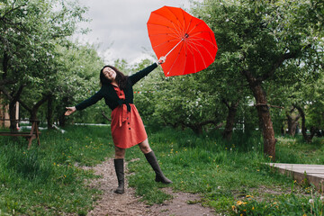 Pretty young woman carrying heart shaped umbrella and looking at camera with smile in the garden. Having fun under the rain