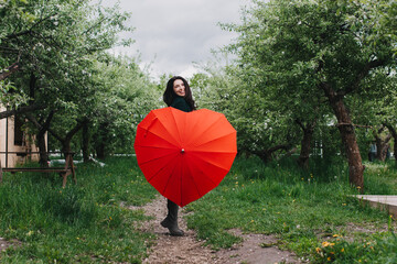 Pretty young woman carrying heart shaped umbrella and looking at camera with smile in the garden. Rain check