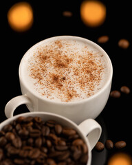 Close up of cup of frothy cappuccino sprinkled with cinnamon and cup with coffee beans in blurred foreground. Objects on black background with spots of light. Vertical format. Soft focus.