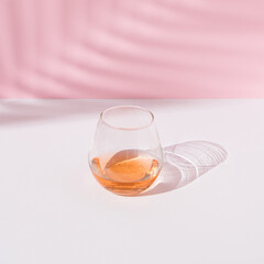 A glass of strong drink in the sun, its shadow and the shadow of a palm tree on a white-pink background