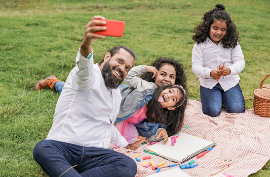 Happy indian family enjoy day outdoor at city park with pic nic and toys while taking a selfie with smartphone - Family, parents and children love
