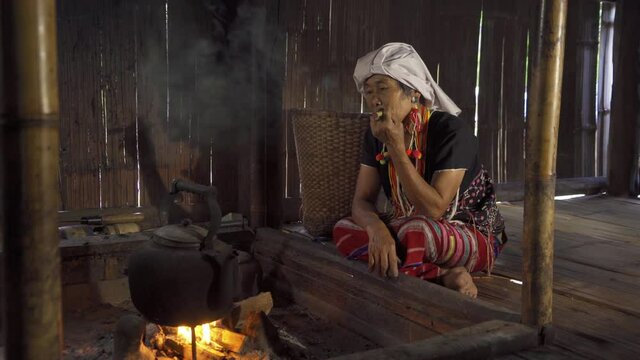 A portrait of karen tribe woman using cigarette to smoke, boiling water by using traditional kettle and fire, water heater at wooden kitchen at local home. People lifestyle.