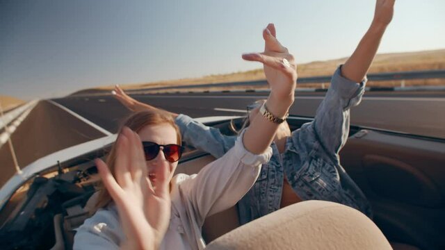 Attractive young women in sunglasses ride convertible with their arms outstretched, dancing, while driving on highway