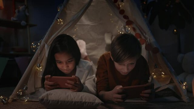 Excited teenagers spending time while playing online games in decorative makeshift hut at home in evening. Girl and boy lying on floor while using smartphones.Concept of leisure.