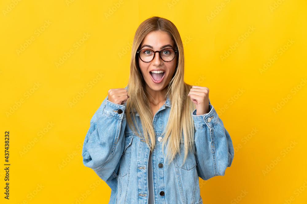 Sticker Young blonde woman isolated on yellow background celebrating a victory in winner position