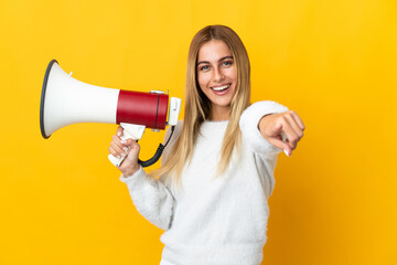 Young blonde woman isolated on yellow background holding a megaphone and smiling while pointing to...