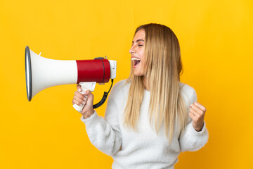 Young blonde woman isolated on yellow background shouting through a megaphone to announce something...