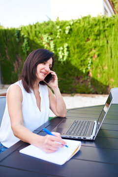 A Caucasian brunette woman teleworking outdoors with her laptop and talking on the mobile phone
