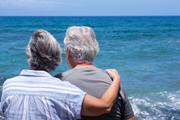 Back view of senior couple sitting on the beach looking at sea. Two retired enjoying summer vacation and freedom