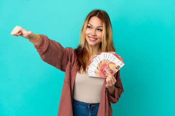 Teenager blonde girl taking a lot of Euros over isolated blue background giving a thumbs up gesture