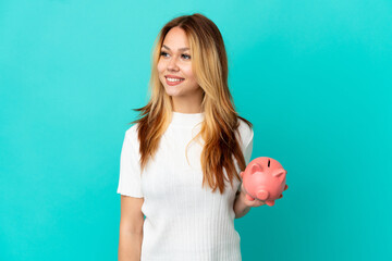 Teenager blonde girl holding a piggybank over isolated blue background looking side
