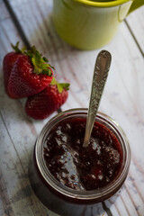 Fotografía cenital de frasco de  mermelada de frutilla con tres frutillas fresca  y una taza.