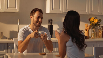 happy man holding cup and looking at brunette girlfriend in kitchen