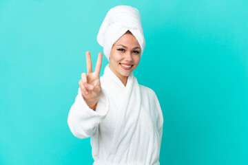 Teenager blonde girl in a bathrobe over isolated blue background smiling and showing victory sign