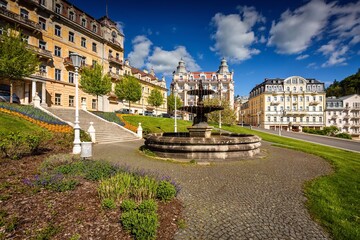 Fototapeta premium Marianske Lazne, Czech Republic - May 30 2021: The baroque water fountain standing on Goethe square. Green lawn, flowers and hotel buildings around. Sunny day with blue sky and clouds in spa city.