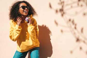 Beautiful black woman with afro curls hairstyle.Smiling hipster model in yellow hoodie. Sexy carefree female posing on the street background near wall in sunglasses. Cheerful and happy