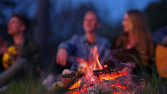 Burning wood in fire on blur background of people. Young friends having a picnic by the campfire at night. Happy friendship.