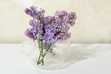 composition of a bouquet of lilacs in a glass teapot on a white table on a beige background