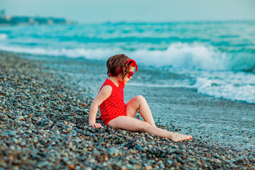 a little girl in a red swimsuit and sunglasses, sitting on the beach in summer