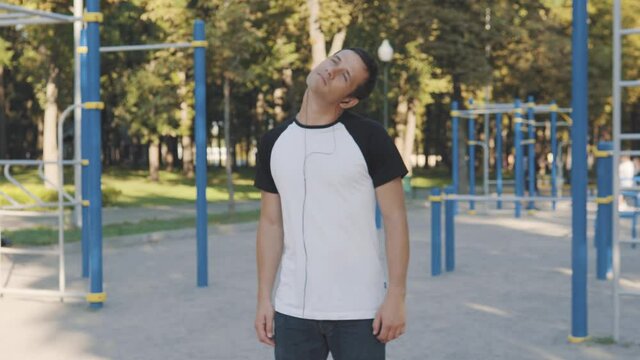 An attractive man rotates his head, kneads his neck near the fitness park on a bright sunny day.
