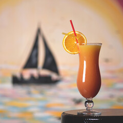 Tropical cocktail drink. Beverage in glass goblet decorated with orange slice and straw. Sailboat on blurred background. Vacation or summer holiday concept. Copy space to left. Soft focus.