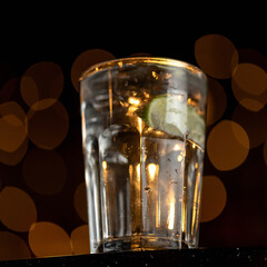 Backlit Glass with drink or beverage at table on black background with bokeh. Soft focus. Square format. Side view. 