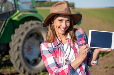 Fototapeta premium Woman farmer in front of tractor showing tablet looking at camera smiling