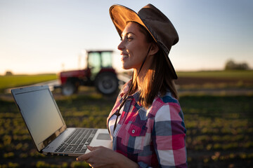 Portrait of young female farmer using laptop in field in front of tractor.