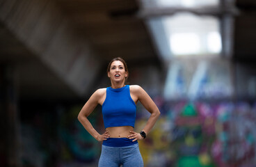 Female athlete standing with hands on hips breathing after workout in urban setting.