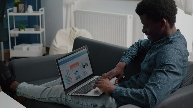 African american man working from home, remote working on laptop computer with online graphs and statistics from living room. Computer user using business internet online web communication job