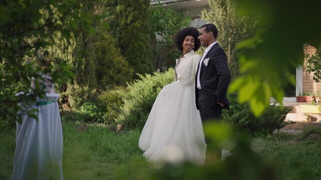Side view wide shot of happy African American couple of newlyweds walking in slow motion in summer garden. Smiling beautiful woman and confident handsome man strolling outdoors on marriage ceremony