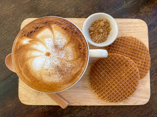 Top view of a cup of cappuccino, Latte art coffee in white cup on wooden board served with two pieces Dutch waffle (Stroopwafel) on the side and brown sugar. 