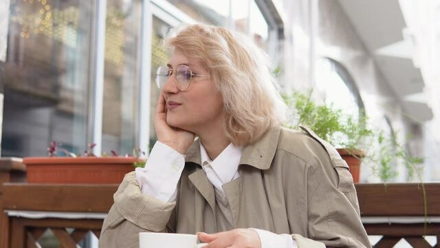 Relaxed woman sitting with a cup of coffee at a table on the terrace. A woman in a trench coat and white blouse picks up a white coffee cup and puts it back on the table. Mindfullness