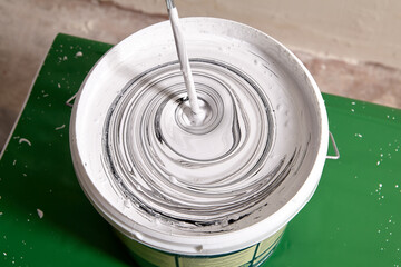 Adding black pigment to white wall paint for mixing in bucket using an electric mixer until gray...