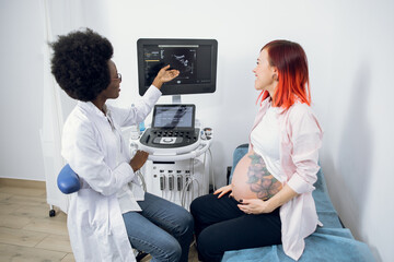 Beautiful young red haired pregnant woman in modern clinic, looking at the screen of ultrasound...