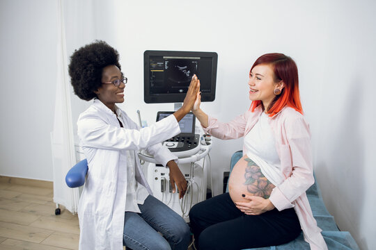 Happy attractive pregnant Caucasian woman patient giving her smiling African female obstetrician a high five, talking about pregnancy after ultrasound scanning