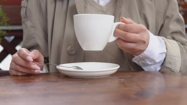 A young woman with a neat manicure in beige and white casual clothes picks up a white porcelain cup of coffee and puts it back on a saucer. Close-up view of hands and cups