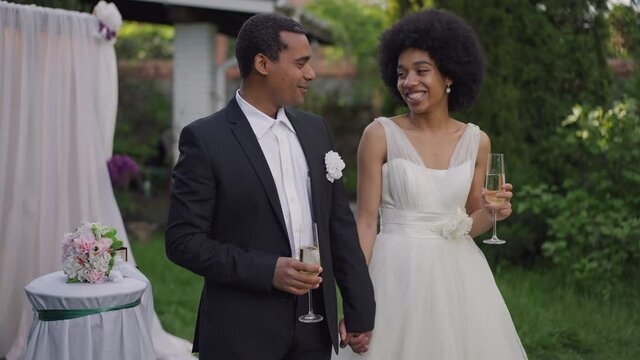 Couple of joyful African American newlyweds clinking champagne glasses toasting to guests on marriage ceremony. Happy bride and groom holding hands standing at wedding altar in spring garden smiling