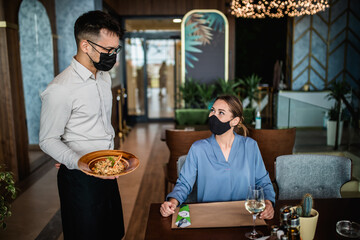 Young waiter with face protective mask serving delicious meal to beautiful woman in restaurant. They are wearing face protective masks due to Coronavirus pandemic.