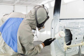 a worker in a gray protective suit are painting car door parts  spray.  hard work. mask and protective shield.