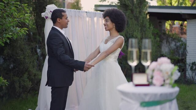 Side view of joyful couple holding hands standing at altar talking in slow motion at sunset. Happy smiling African American bride and groom chatting outdoors. Happiness and marriage