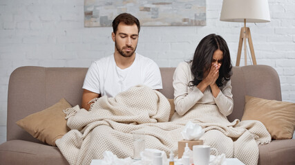 Fototapeta premium sick woman sneezing near man and bottles with pills on coffee table