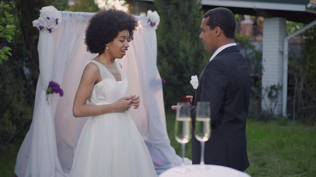 Medium shot side view of young African American man in wedding suit opening ring box as excited woman in wedding dress with toothy smile holding hands at chest. Happy couple at altar outdoors