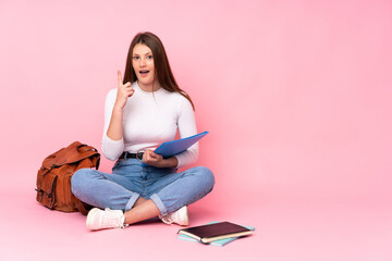 Teenager caucasian student girl sitting on the floor isolated on pink background thinking an idea pointing the finger up